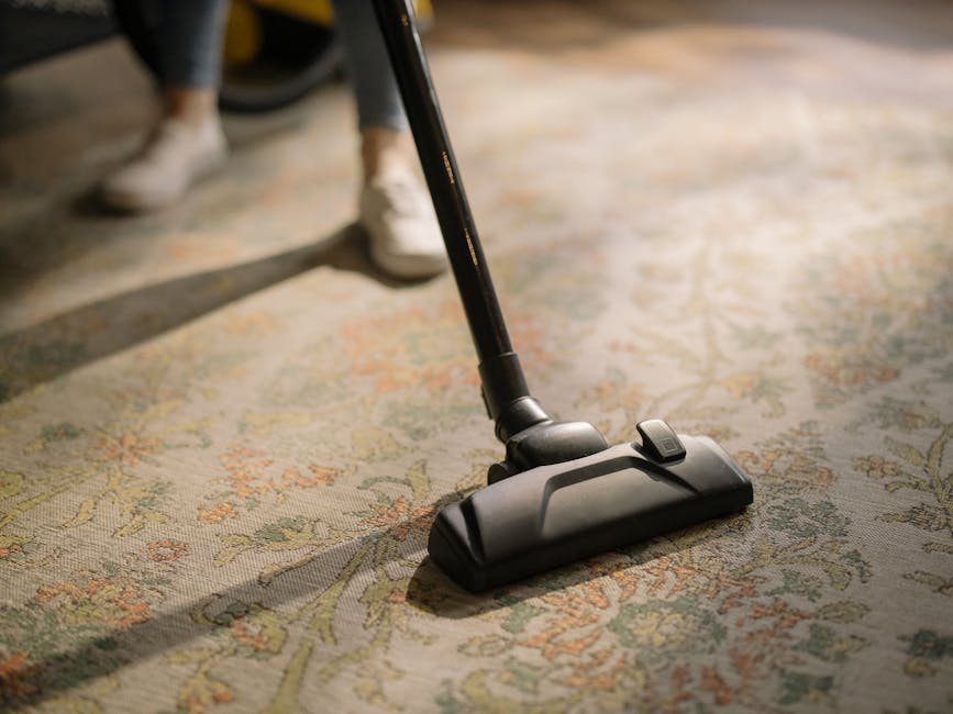 Close-up of a person using a red handheld vacuum cleaner to clean a wooden floor in a domestic setting, with a white chest of drawers and a beige curtain in the background. The vacuum is positioned on